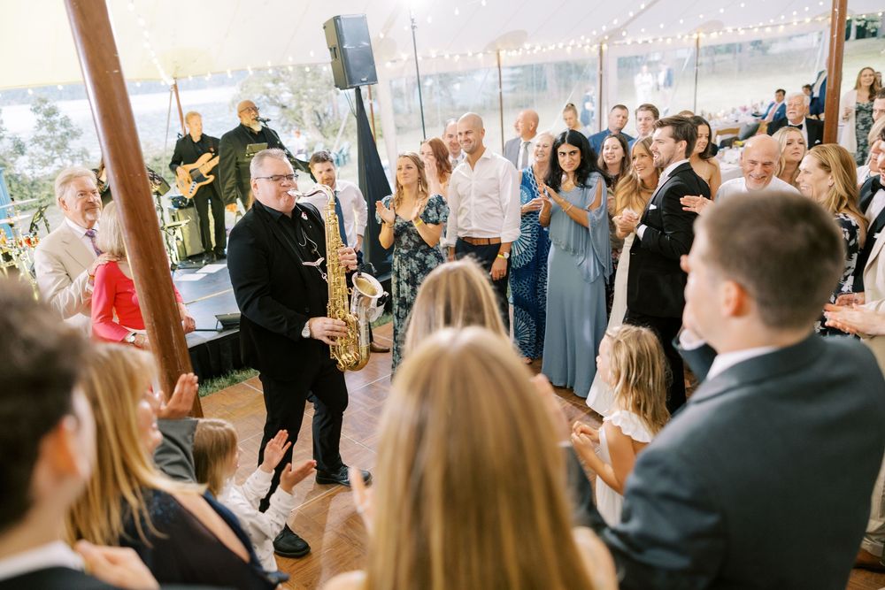 Saxophone musician of Funk Evolution performs at an outdoor wedding reception surrounded by guests in formal attire at Silver Bay YMCA.