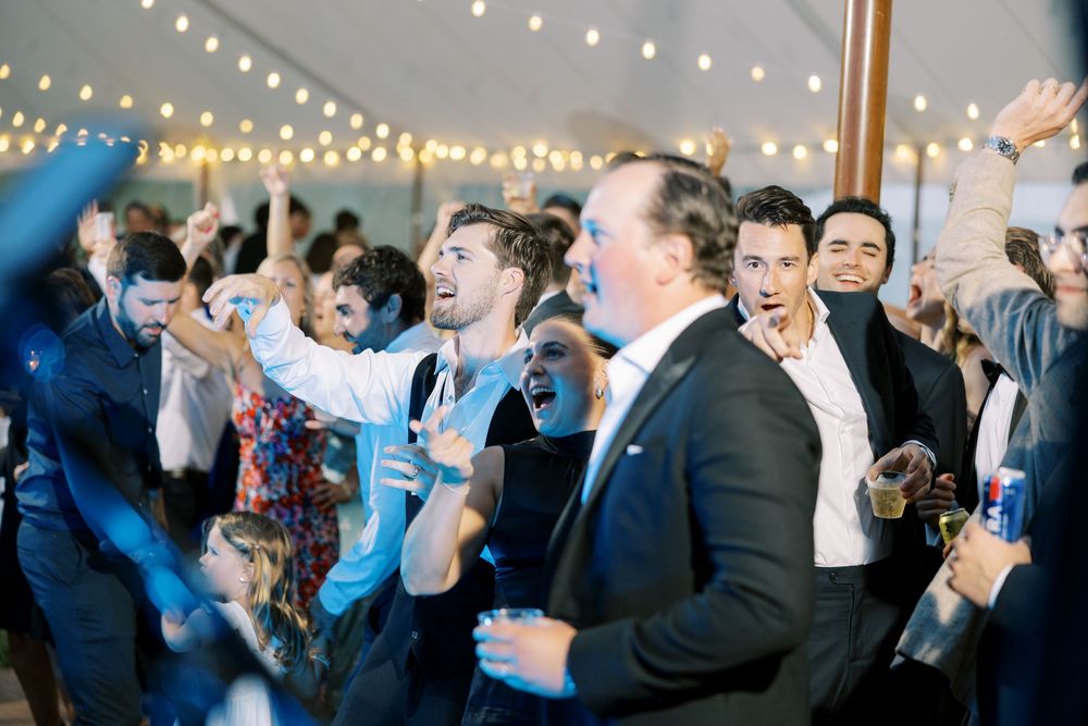 Wedding reception guests dance enthusiastically under twinkling string lights during a lively celebration moment.