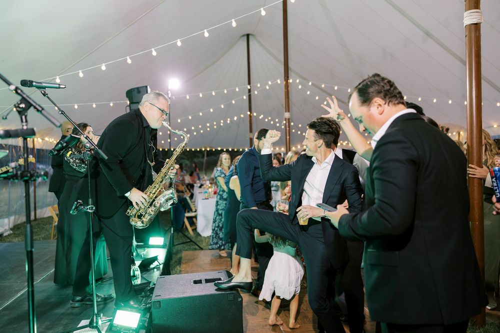 A lively dance party under a tented reception area with string lights glowing overhead and a live band performing.