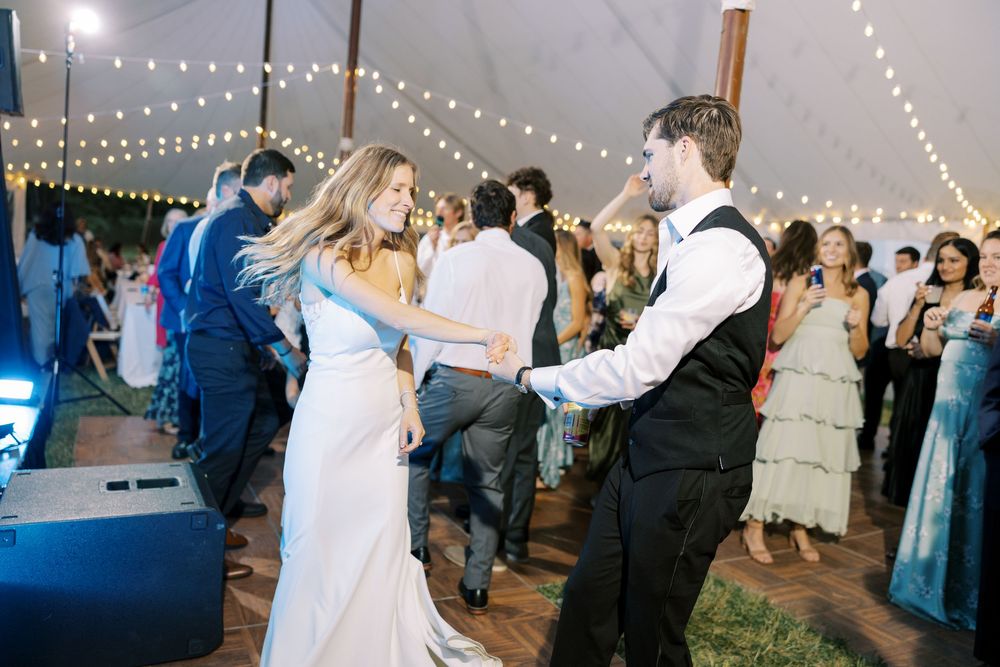 Reception guests dance under string lights in an outdoor tent at an evening wedding celebration.