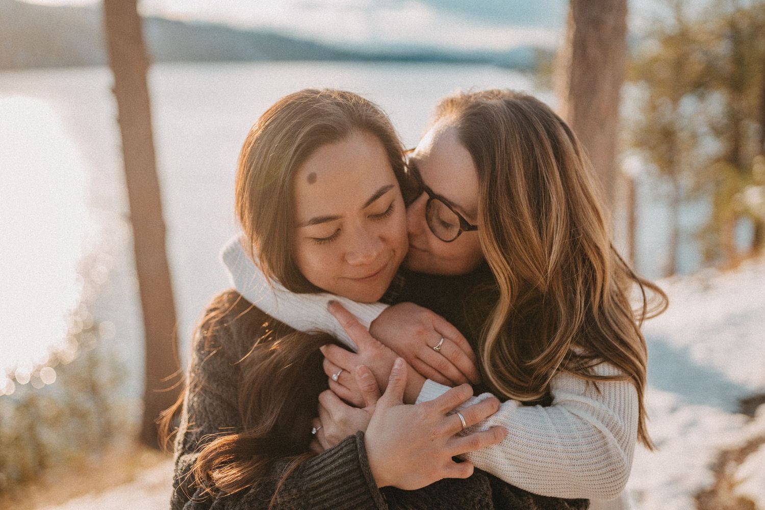 Two people embracing outdoors on a sunny winter day with snow and mountain scenery in the background.
