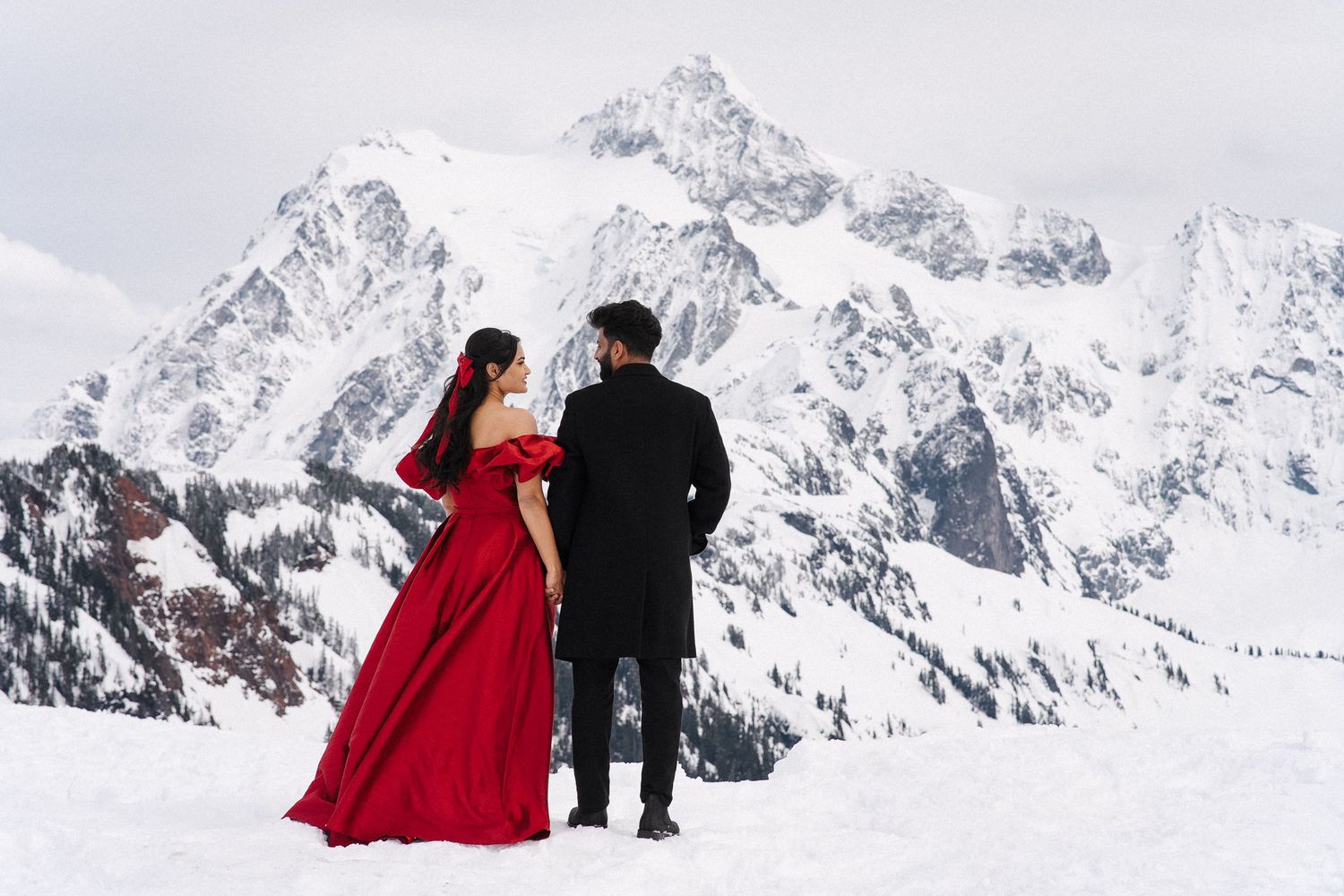 Two people stand together in formal attire against a snowy mountain backdrop, with one wearing a dramatic red gown.