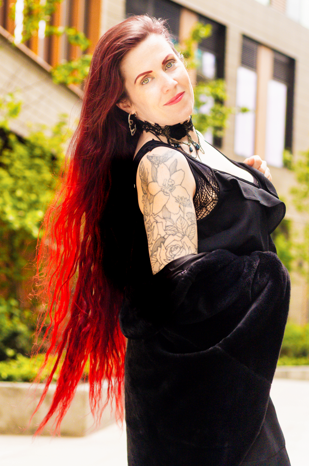 A fashion portrait showcasing long red hair, black dress and floral arm tattoo against urban backdrop.