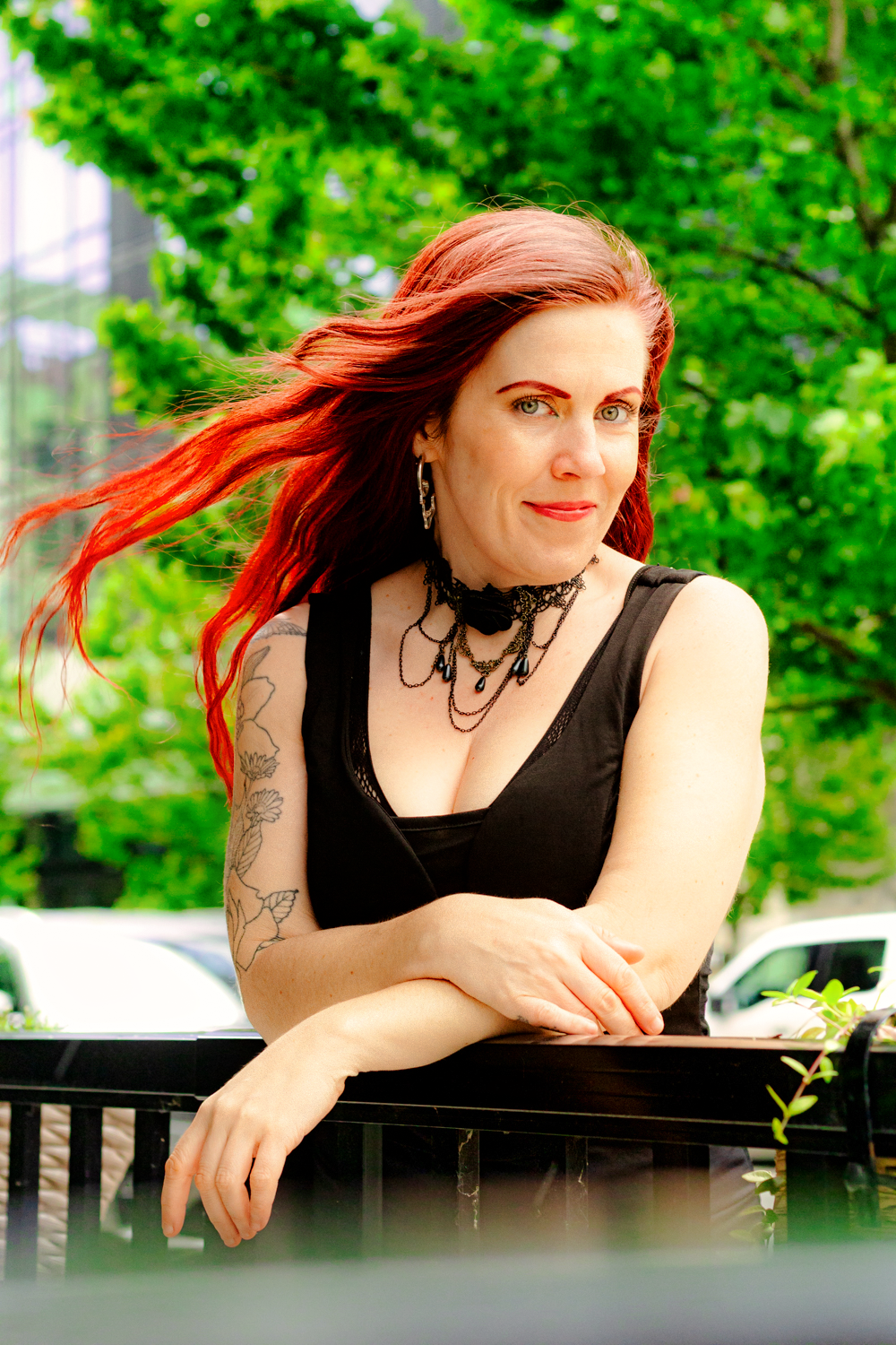 A person in black attire with flowing red hair leans on a railing against green foliage.