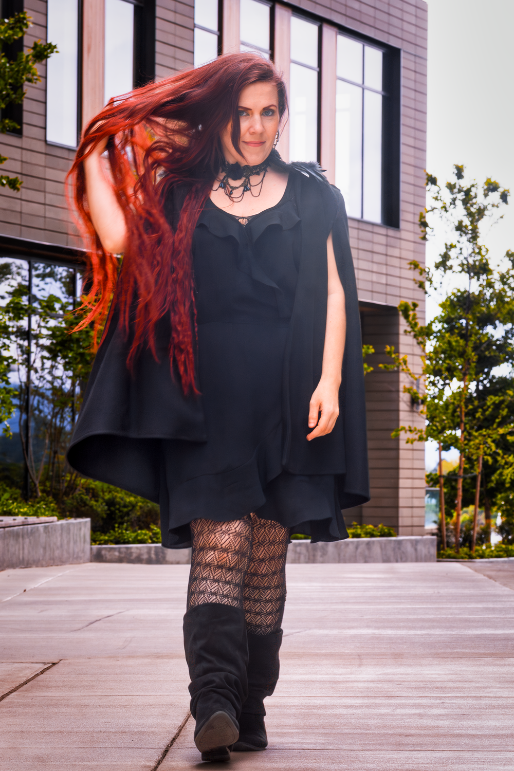 A gothic-styled outfit featuring a black cape dress, patterned tights and boots against an urban backdrop.