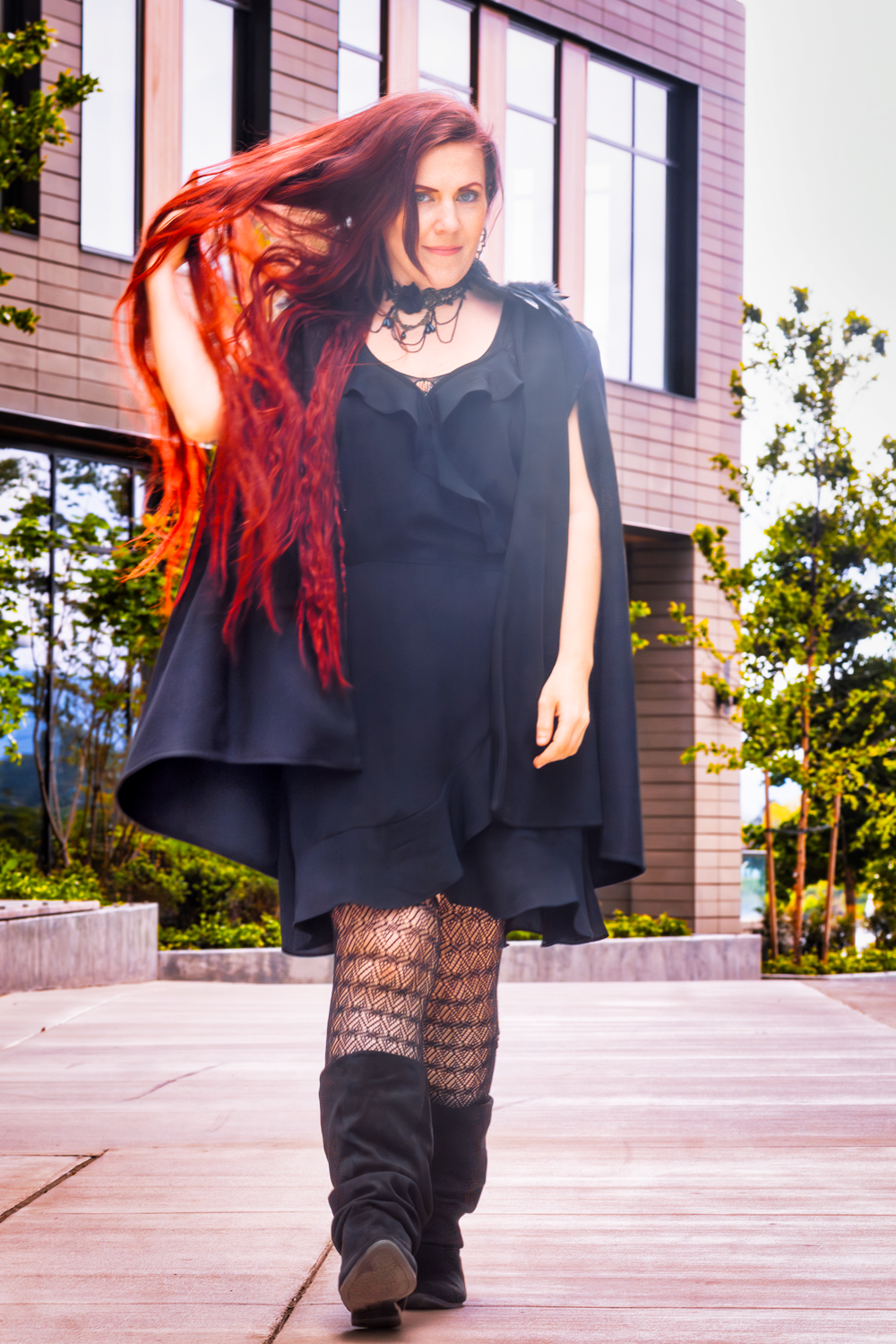 A model with vibrant red hair poses outdoors wearing a black cape dress and patterned tights.