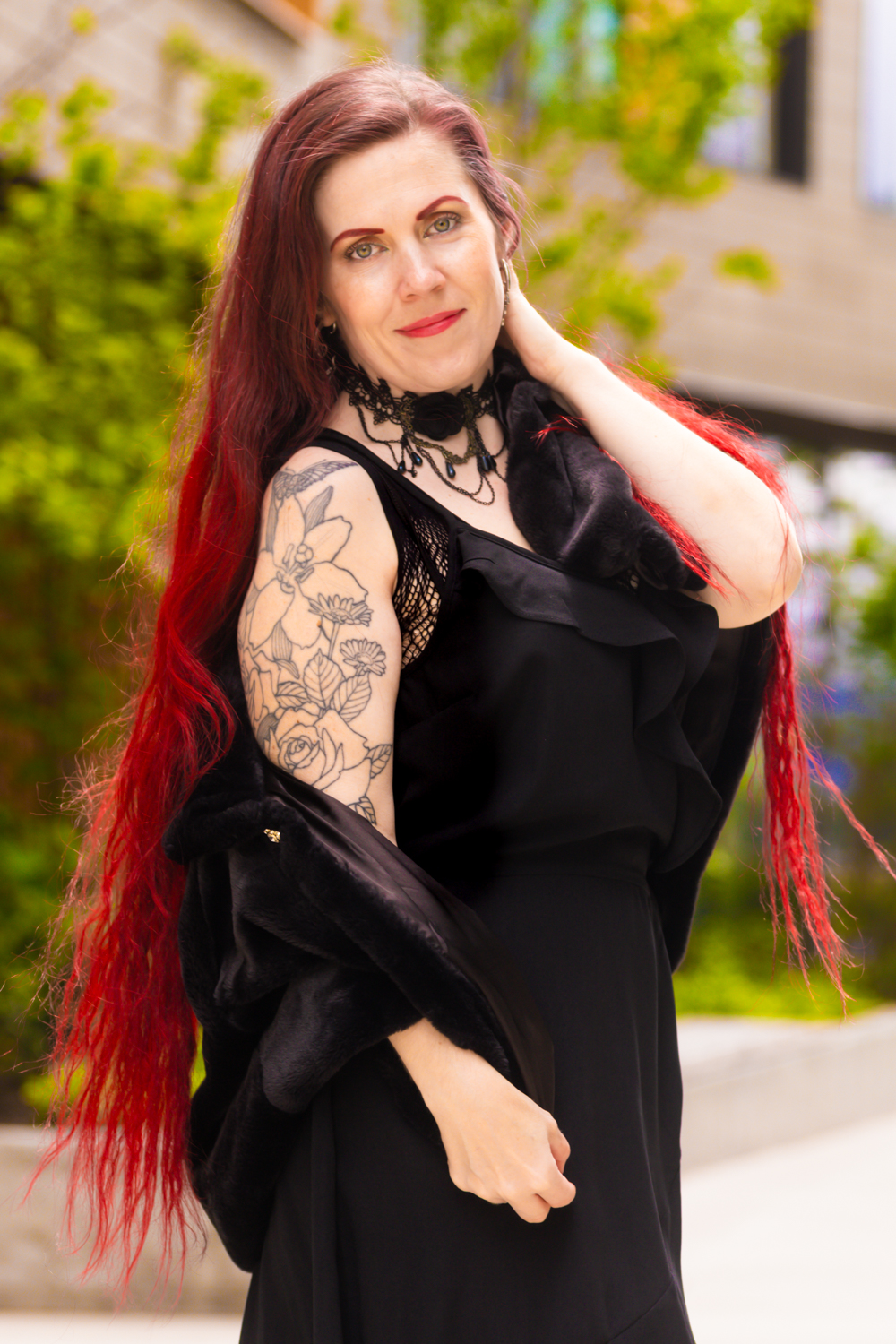 A person in a black dress with bright red hair and arm tattoos poses against a blurred green background.