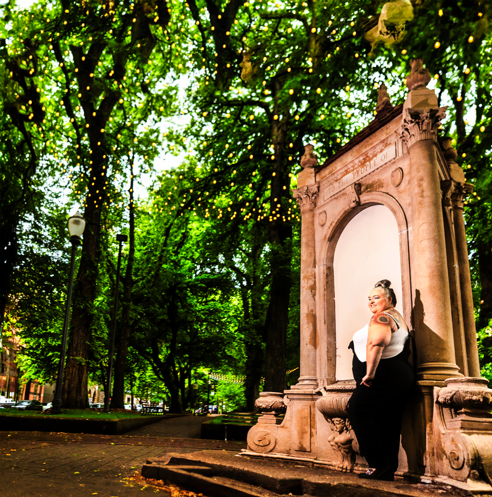 A Gothic figure poses dramatically against an ornate stone archway in a lush green park setting.