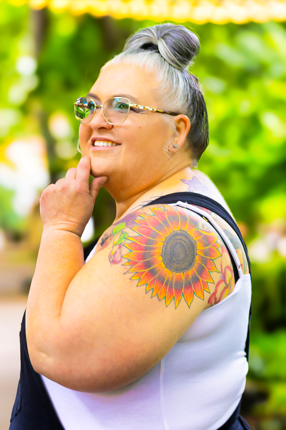 A person with gray hair in a bun and glasses smiles while showing off a vibrant sunflower shoulder tattoo outdoors.