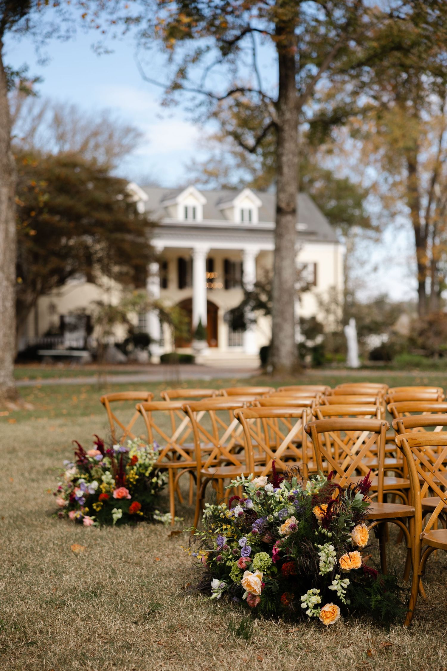 Wooden cross-back chairs arranged for an outdoor wedding ceremony with lush floral arrangements along the aisle.