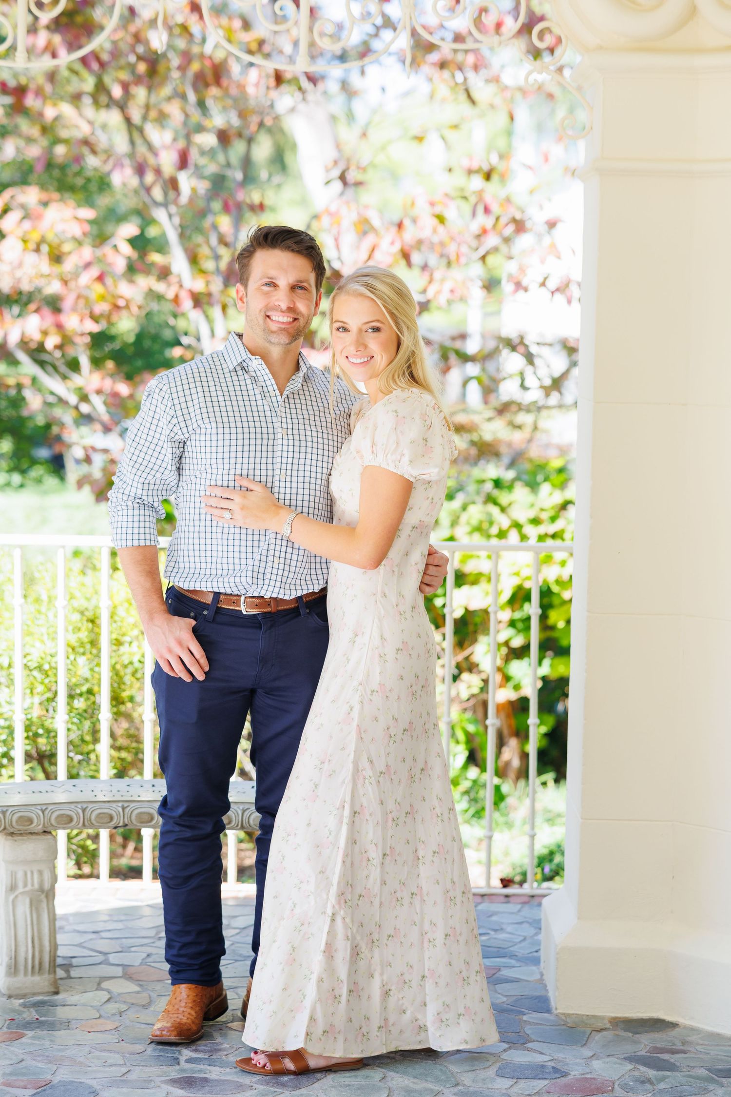 A spring engagement photo in Flippen Park with pink blossoms overhead features a couple in a blue checkered shirt and white lace dress.