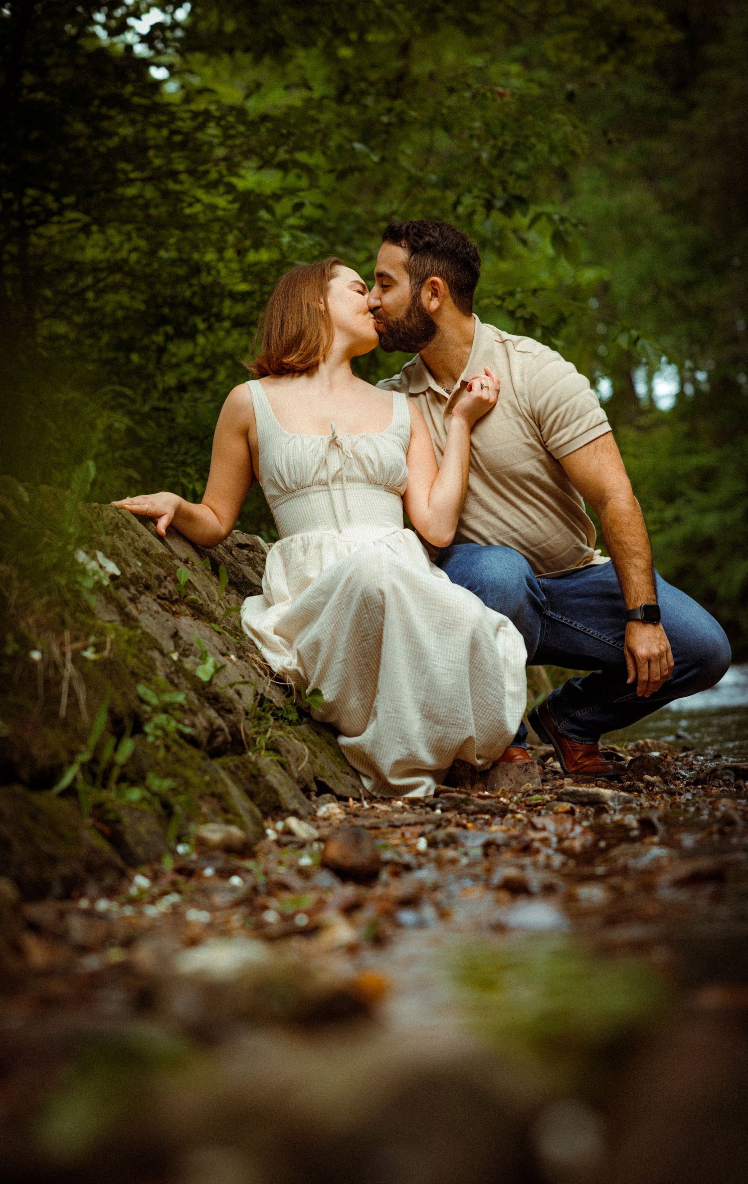 Romantic couple share intimate moment by stream in forest setting with soft natural lighting and autumn leaves.