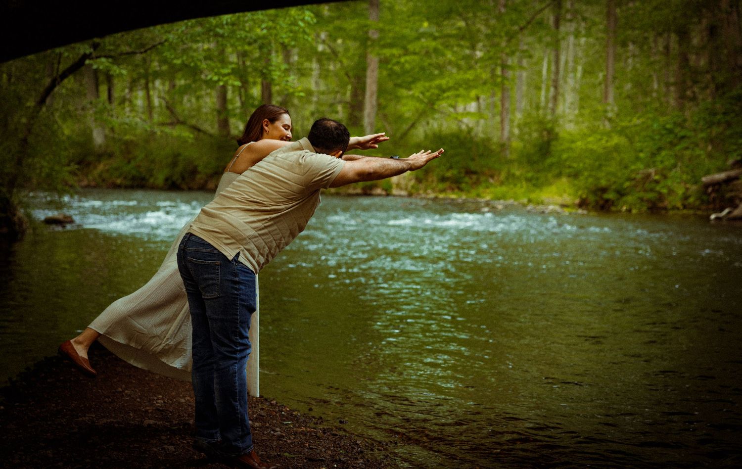 A couple embraces at the edge of a scenic forest stream in a romantic nature setting.
