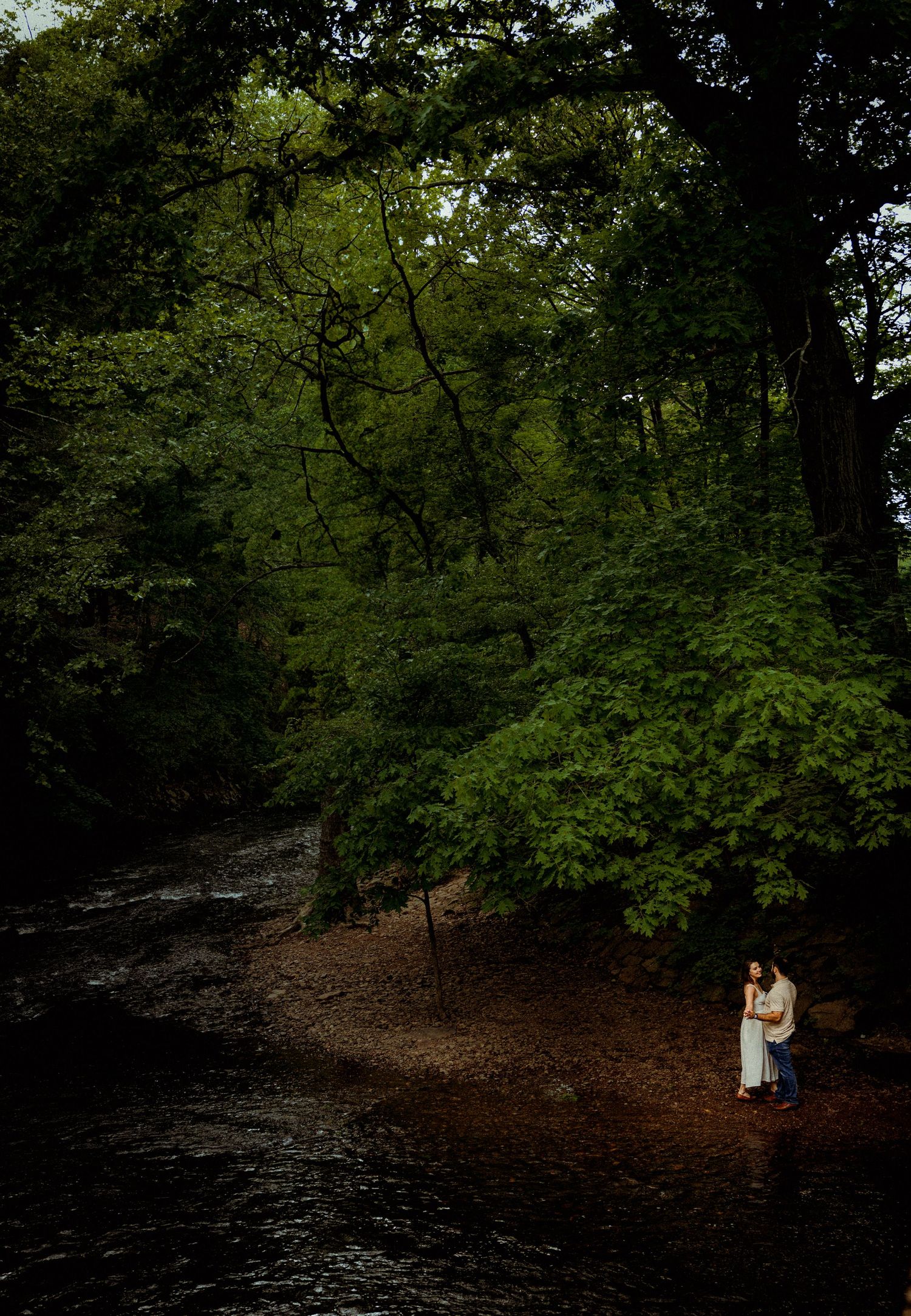 A romantic couple stands by the riverbank in a lush green forest setting.