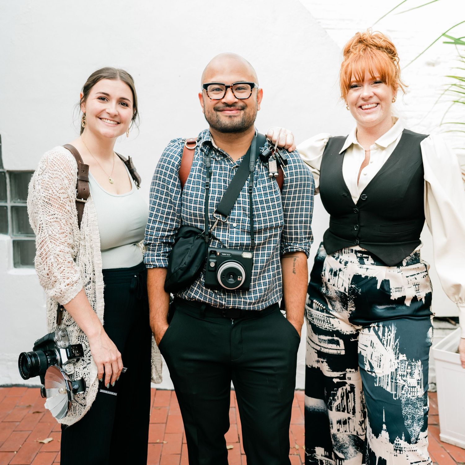 Three photographers with professional cameras stand together smiling outdoors in casual professional attire.