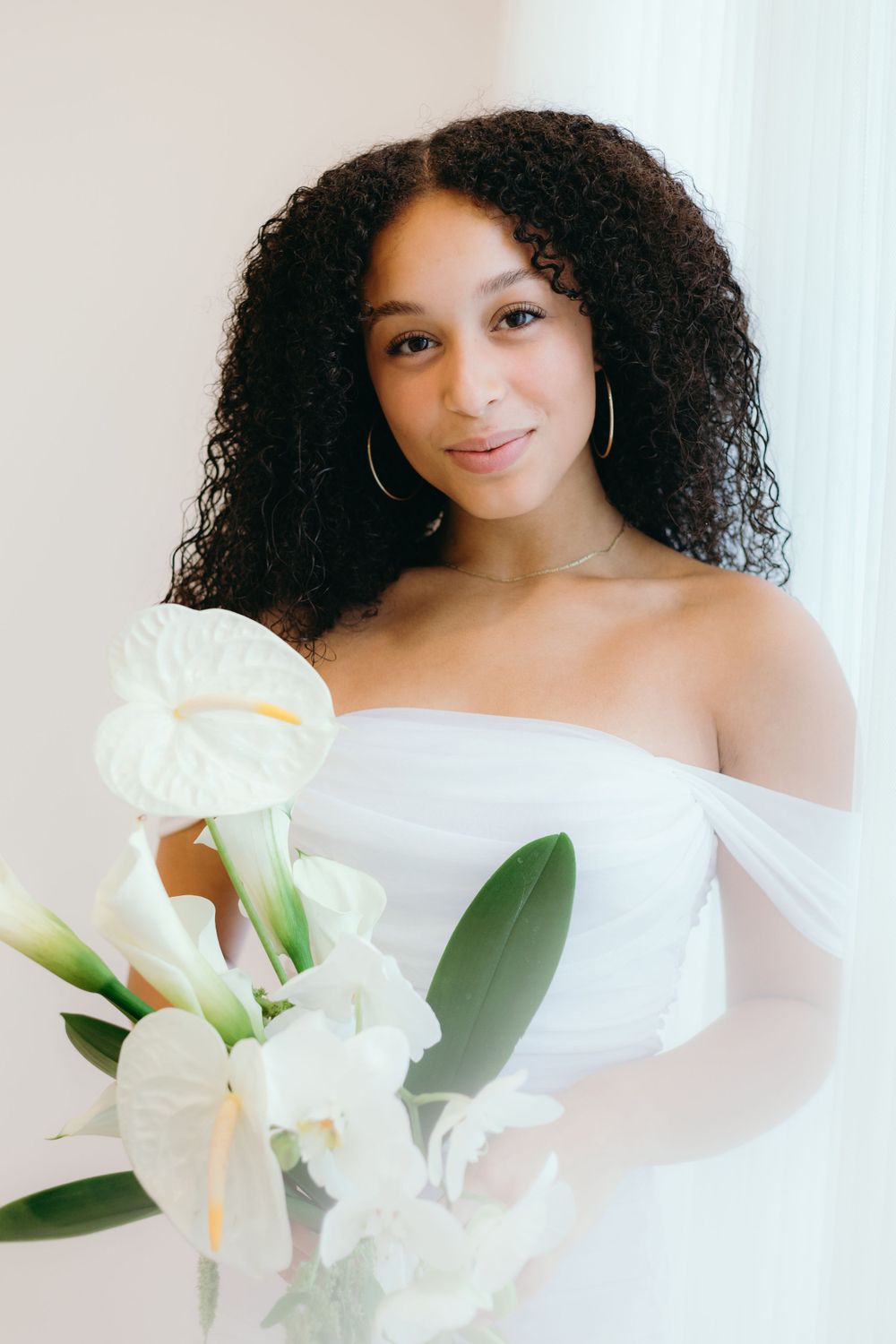 A bride in a white off-shoulder dress holds elegant white lilies while posing against a light background.