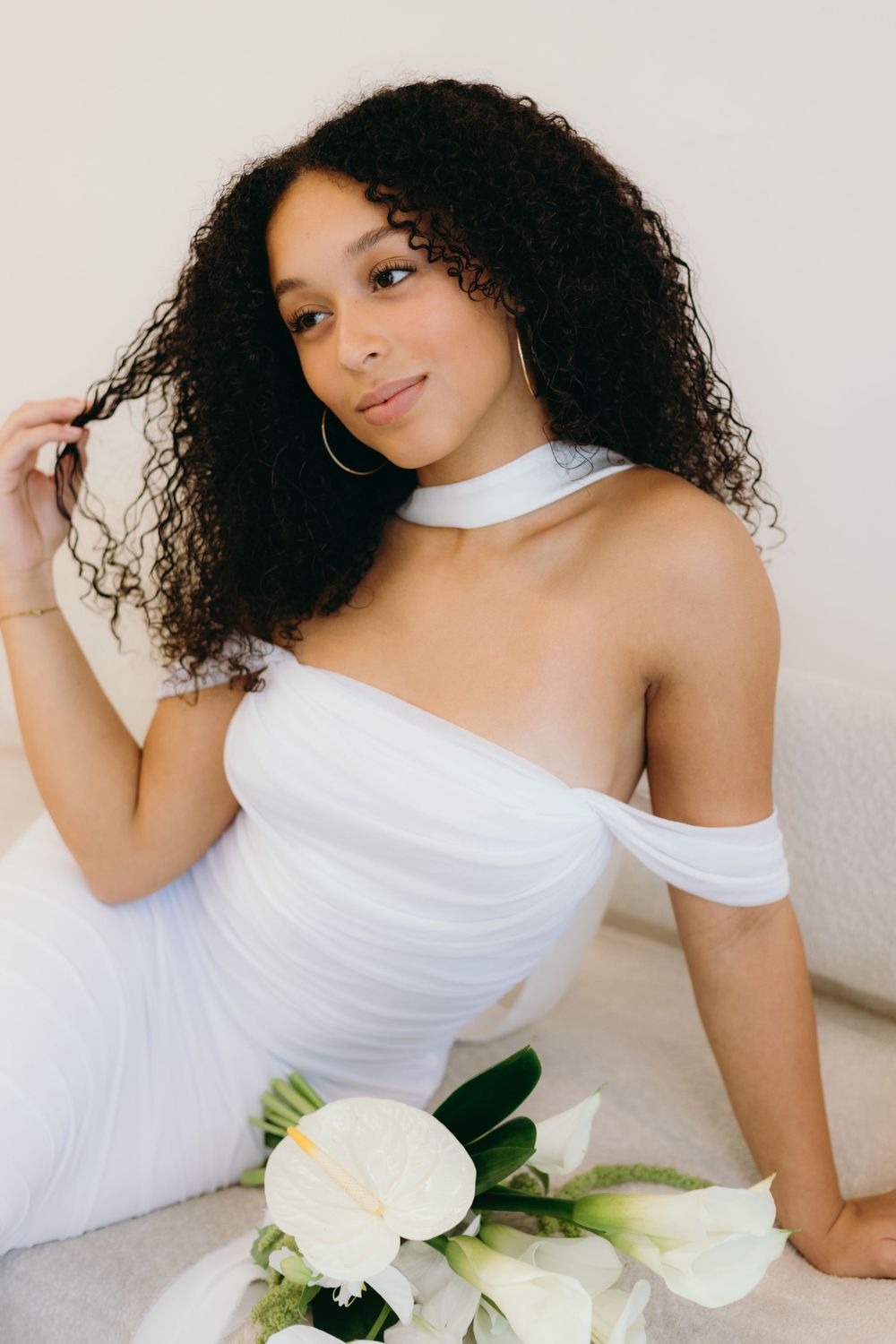 A bride in an elegant off-shoulder white dress poses with calla lilies against a light background.