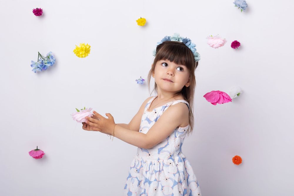 Child in floral dress plays with colorful paper butterflies against a white wall background.