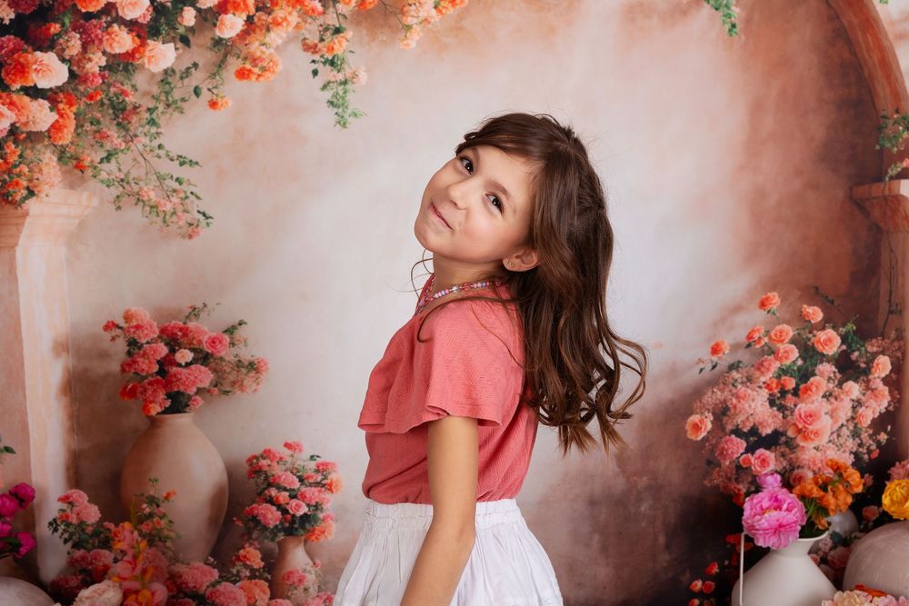 A young figure poses in a pink and white dress against a romantic floral backdrop with coral and pink roses.