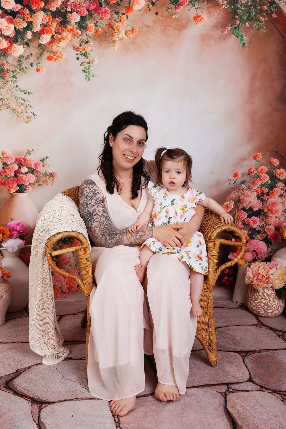 Professional studio photo with floral decor featuring two people seated on wicker chairs against a pink backdrop.