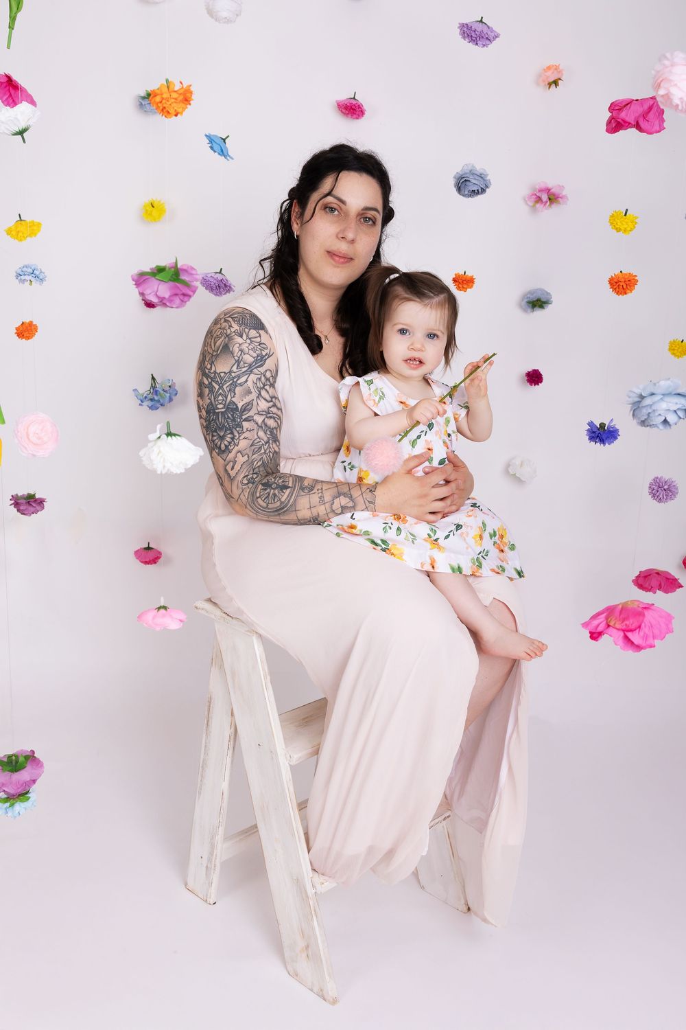 Person in flowy beige dress sits on white chair against colorful butterfly backdrop during portrait session.