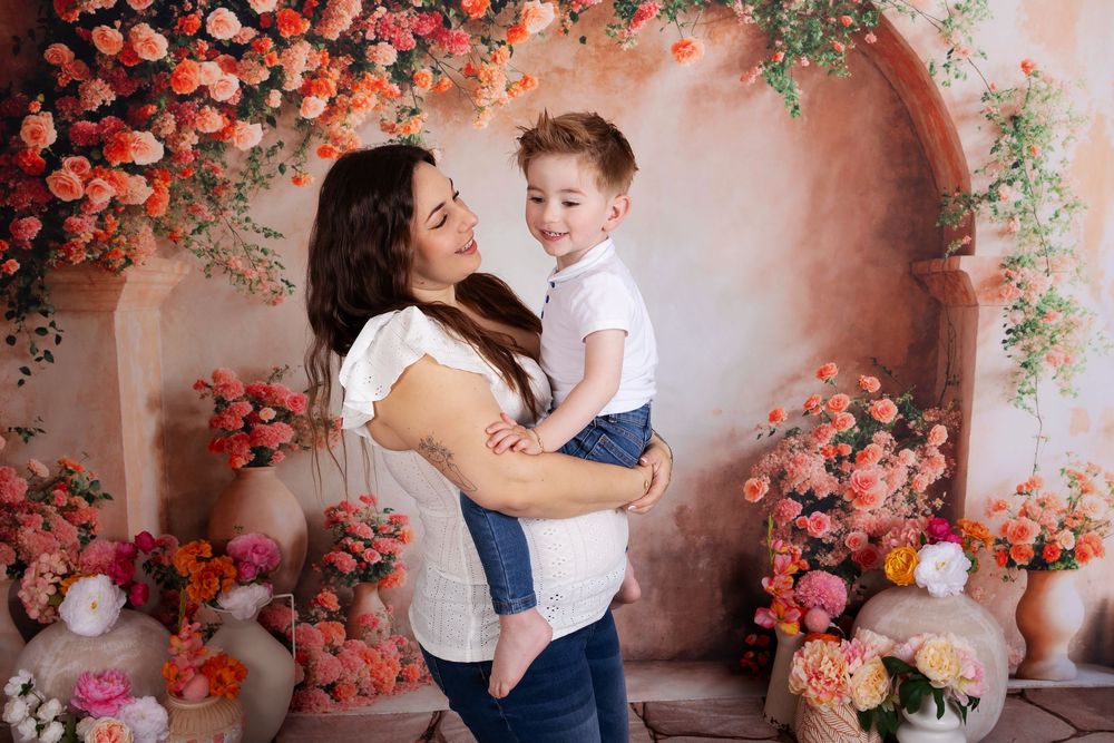 A tender moment captured against a romantic backdrop of coral roses and floral archways in a soft, dreamy studio setting.