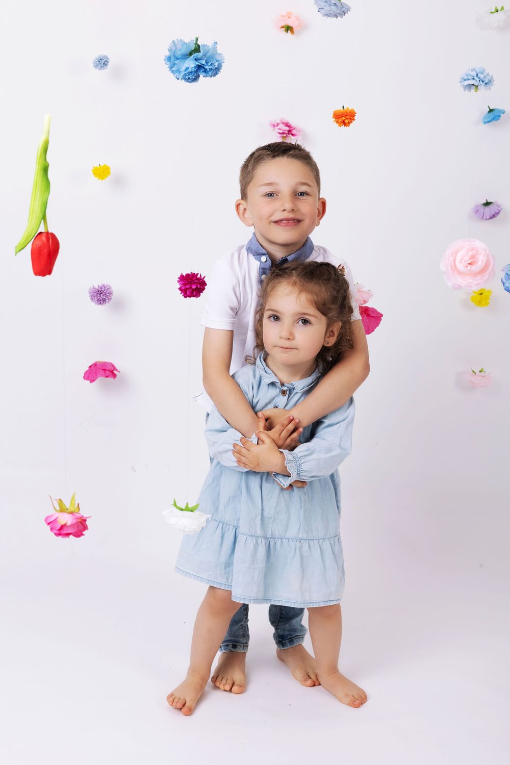 Two young siblings embrace while standing against a white backdrop decorated with colorful paper flowers.