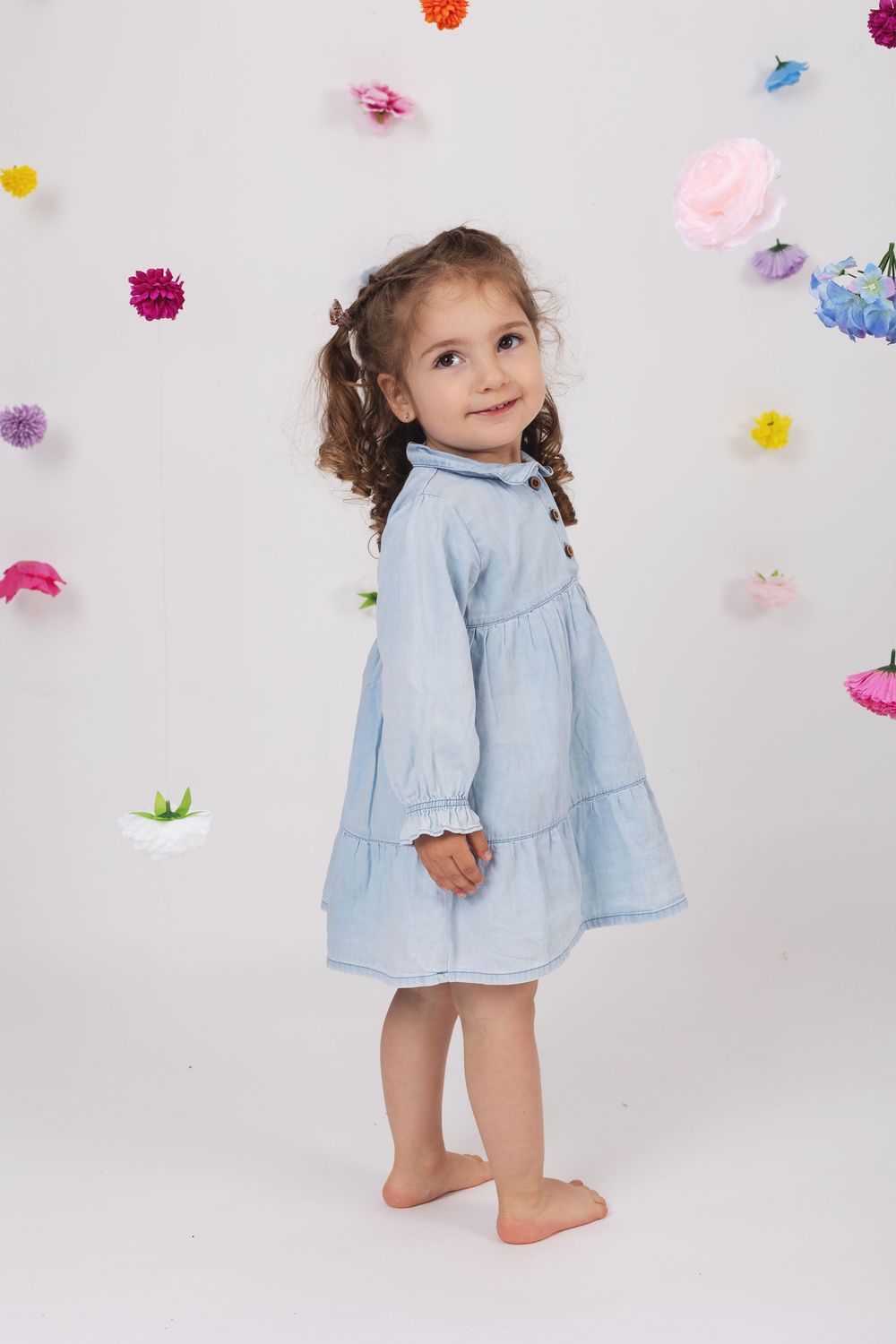 Small child wearing light blue tiered dress stands barefoot against white background with colorful floral decorations.