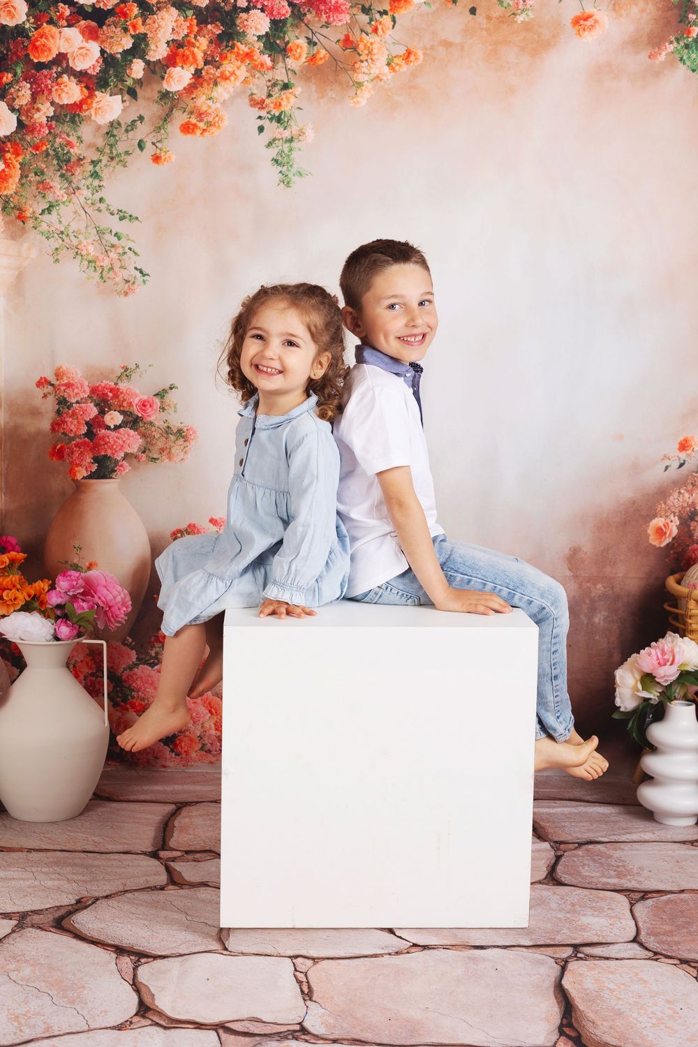 A portrait setup with two young children sitting on a white cube against a floral backdrop with coral roses and greenery.