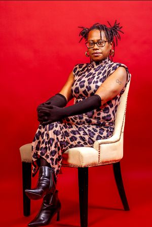 Stylish portrait against red backdrop featuring a leopard print dress, black boots and gloves on an elegant upholstered chair.