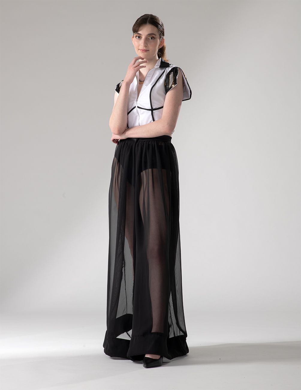 Fashion model wearing white collared top with black trim and sheer black maxi skirt in studio setting.