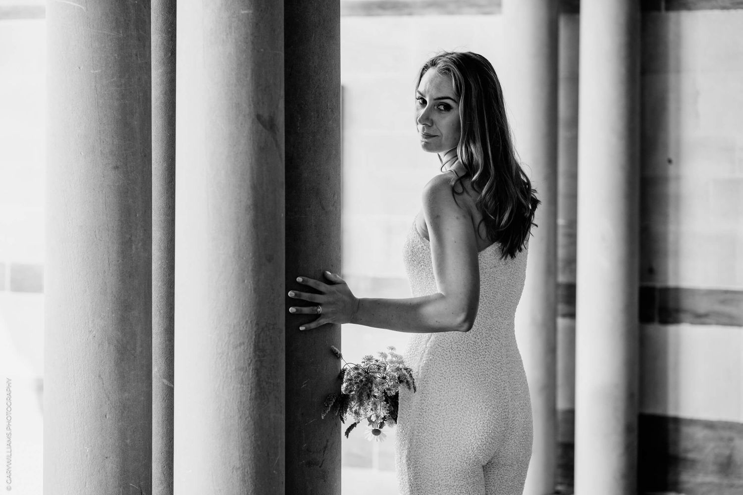 Black and white photo shows an elegant bride in a fitted white gown holding a bouquet by classical columns.