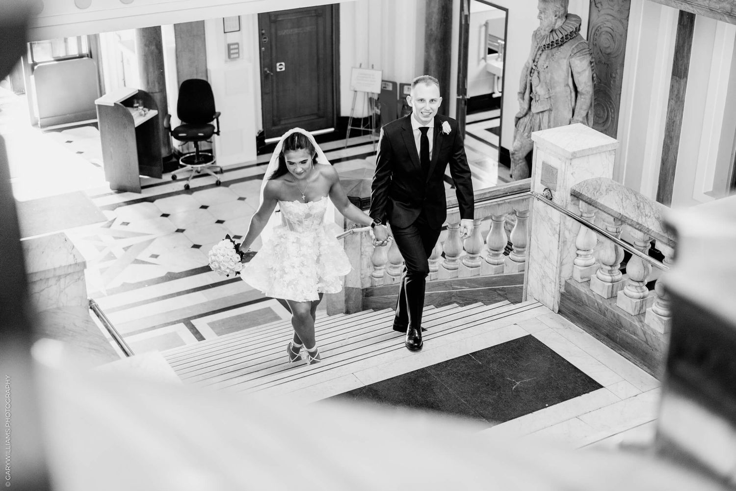 A black and white wedding photo of two people walking down marble stairs in a building corridor together.