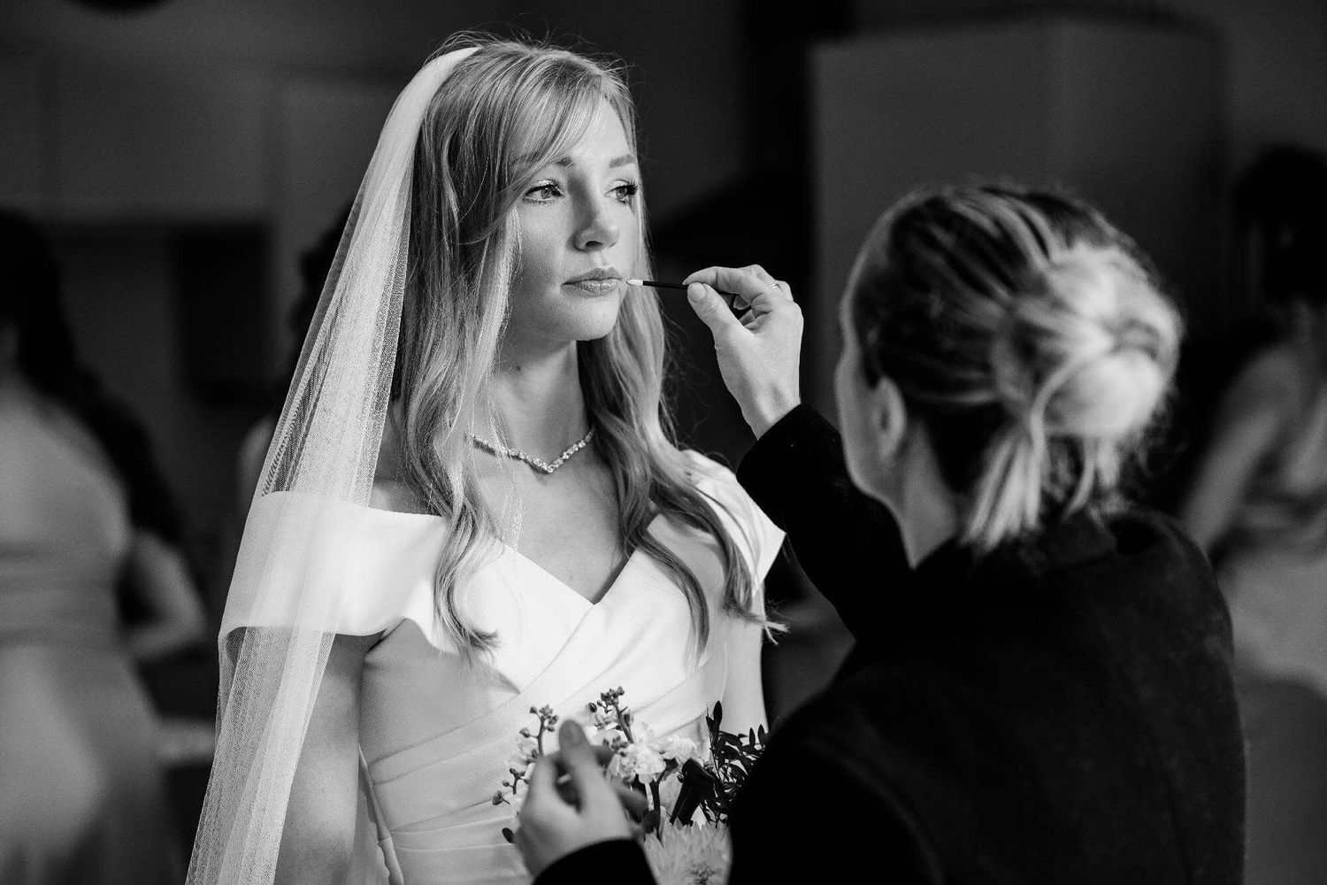 A bride in a white wedding dress and veil gets her makeup touched up by a stylist in a black and white photograph.