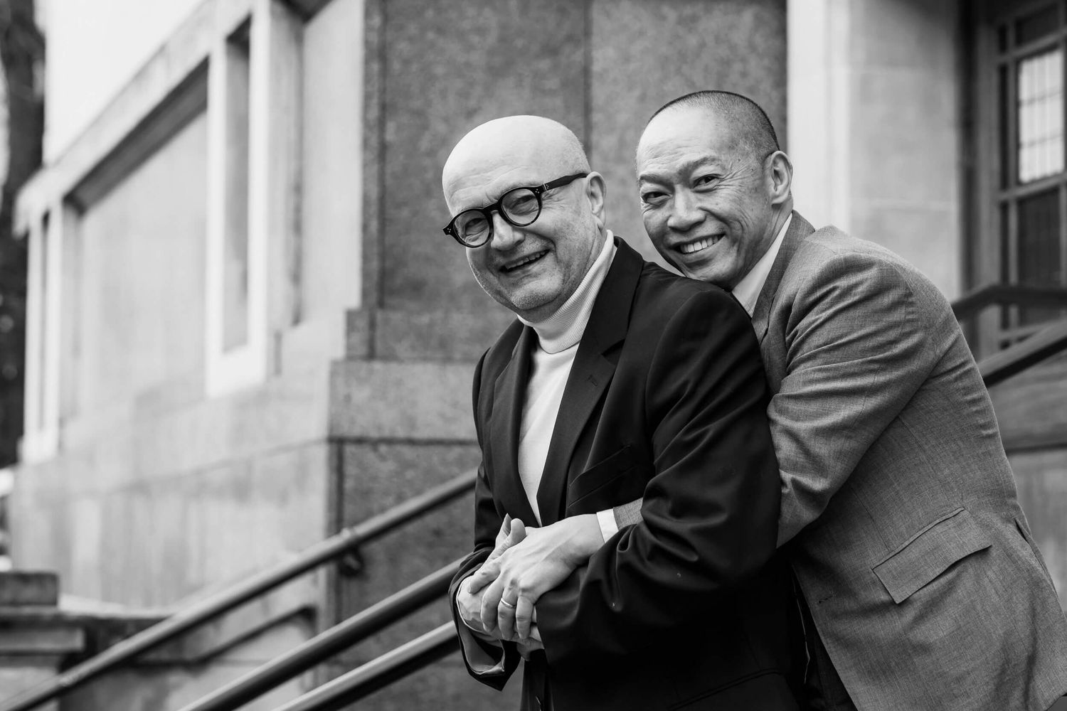 A vintage black and white photograph shows two men in suits smiling together on stone steps outside a building.