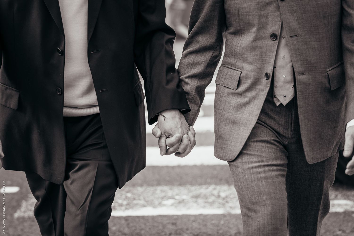 Black and white photo showing two people holding hands while wearing formal suits.