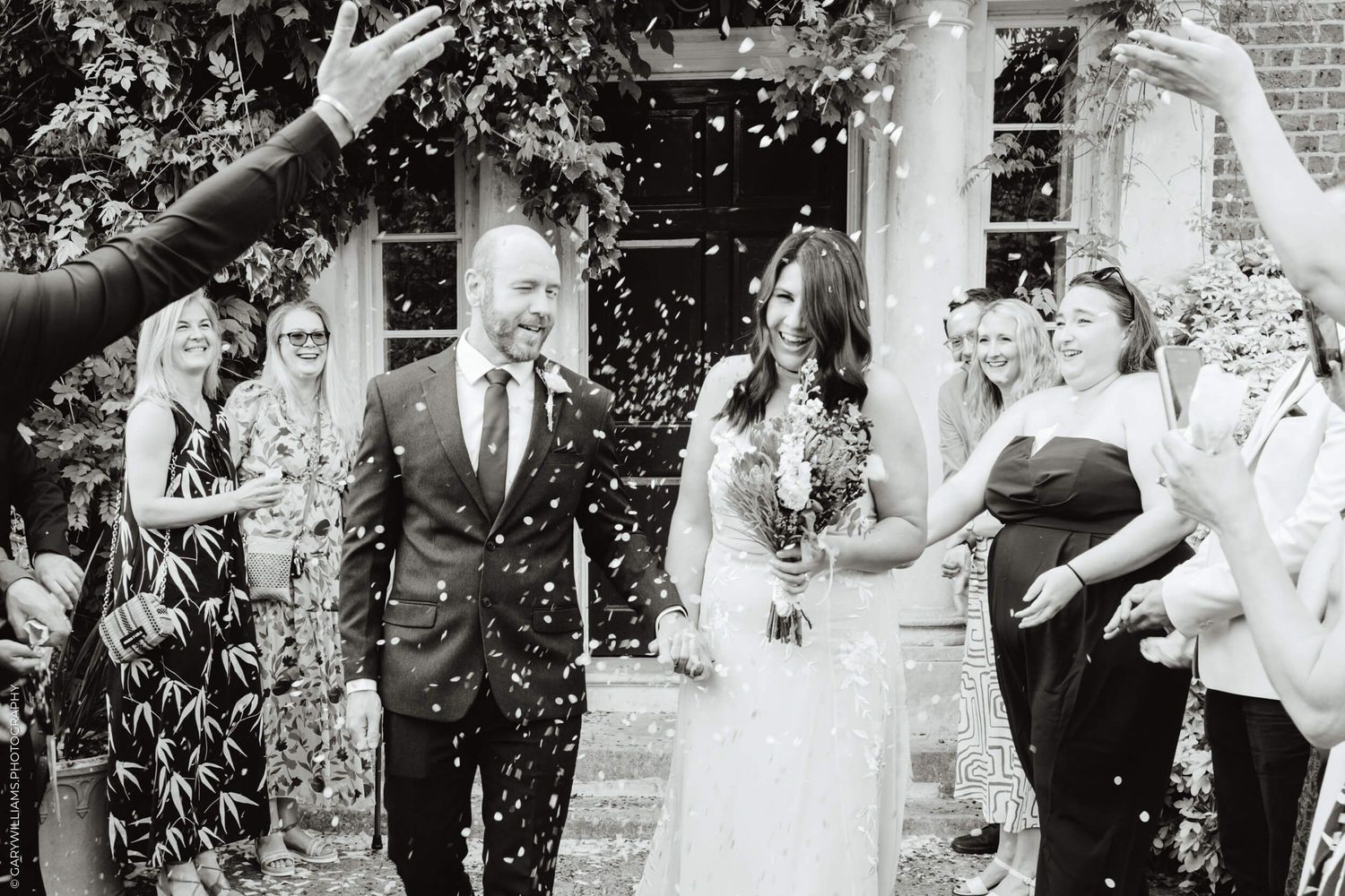 A joyful wedding celebration with guests throwing confetti outside a brick building in black and white photography.
