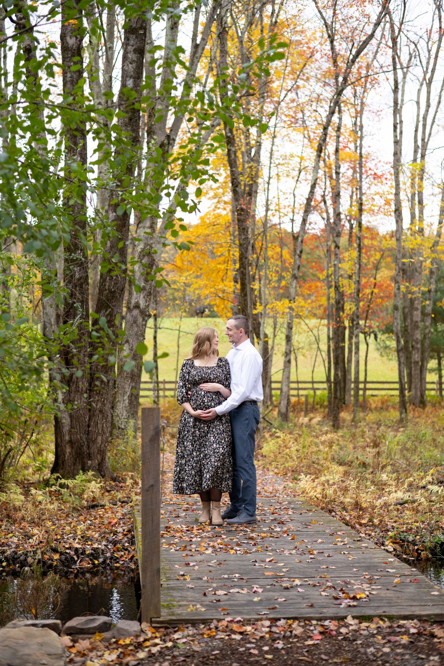 A couple stands together on a wooden bridge surrounded by autumn trees with golden foliage and fallen leaves.