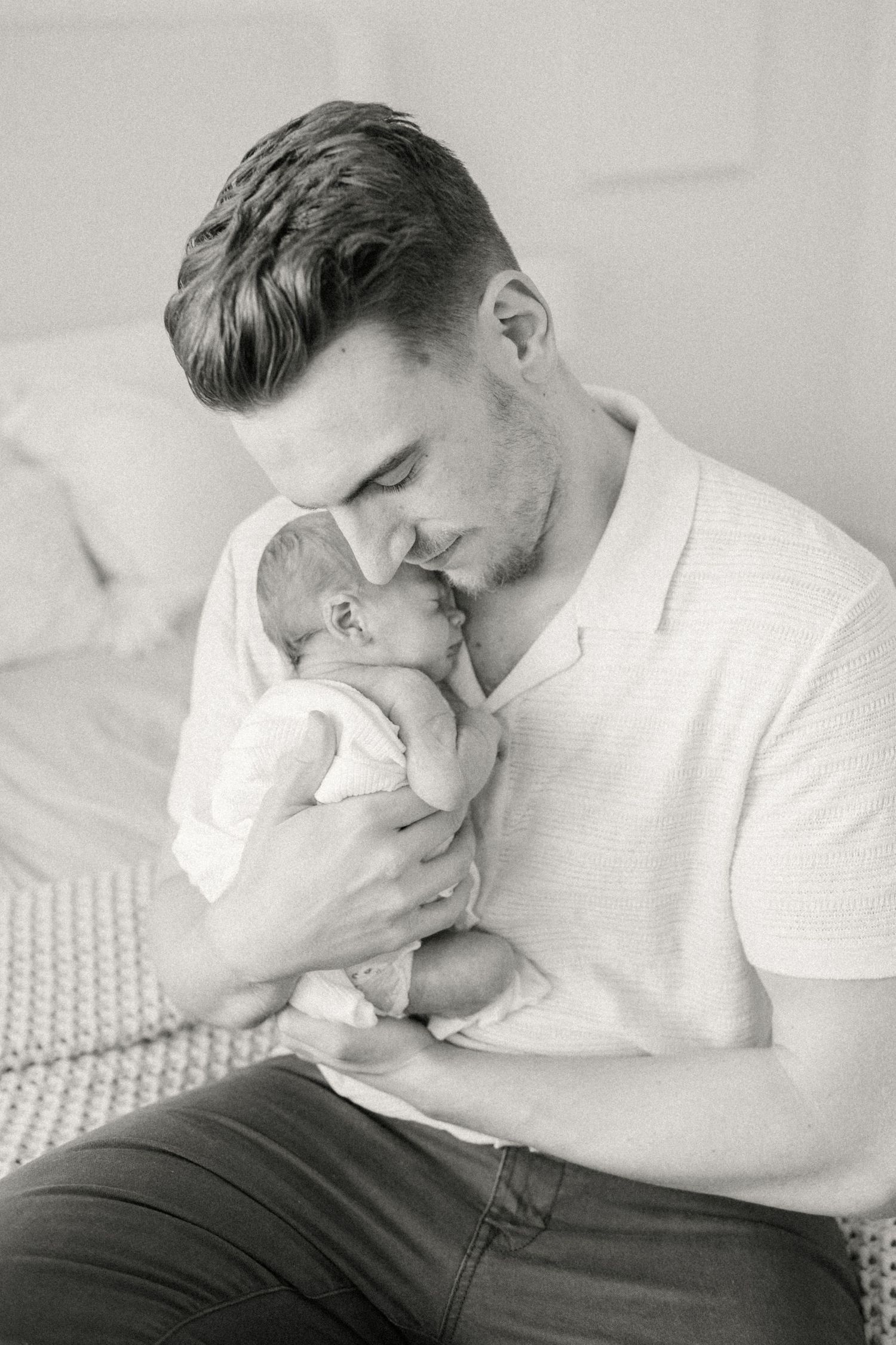 A tender black and white portrait of a parent cradling their newborn baby while sitting on a bed.