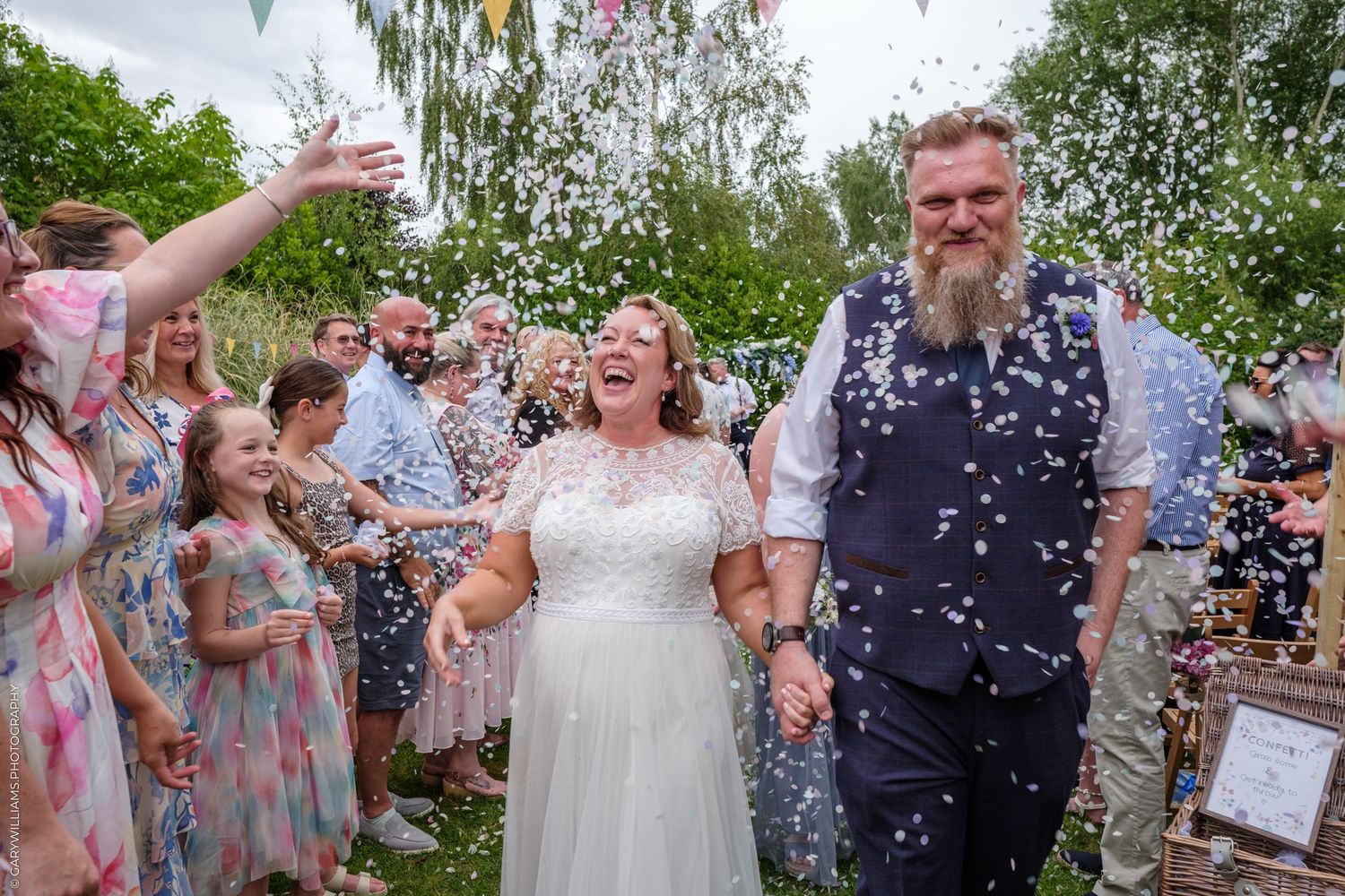 Wedding guests celebrate with confetti as newly married couple walks down garden path during outdoor ceremony.