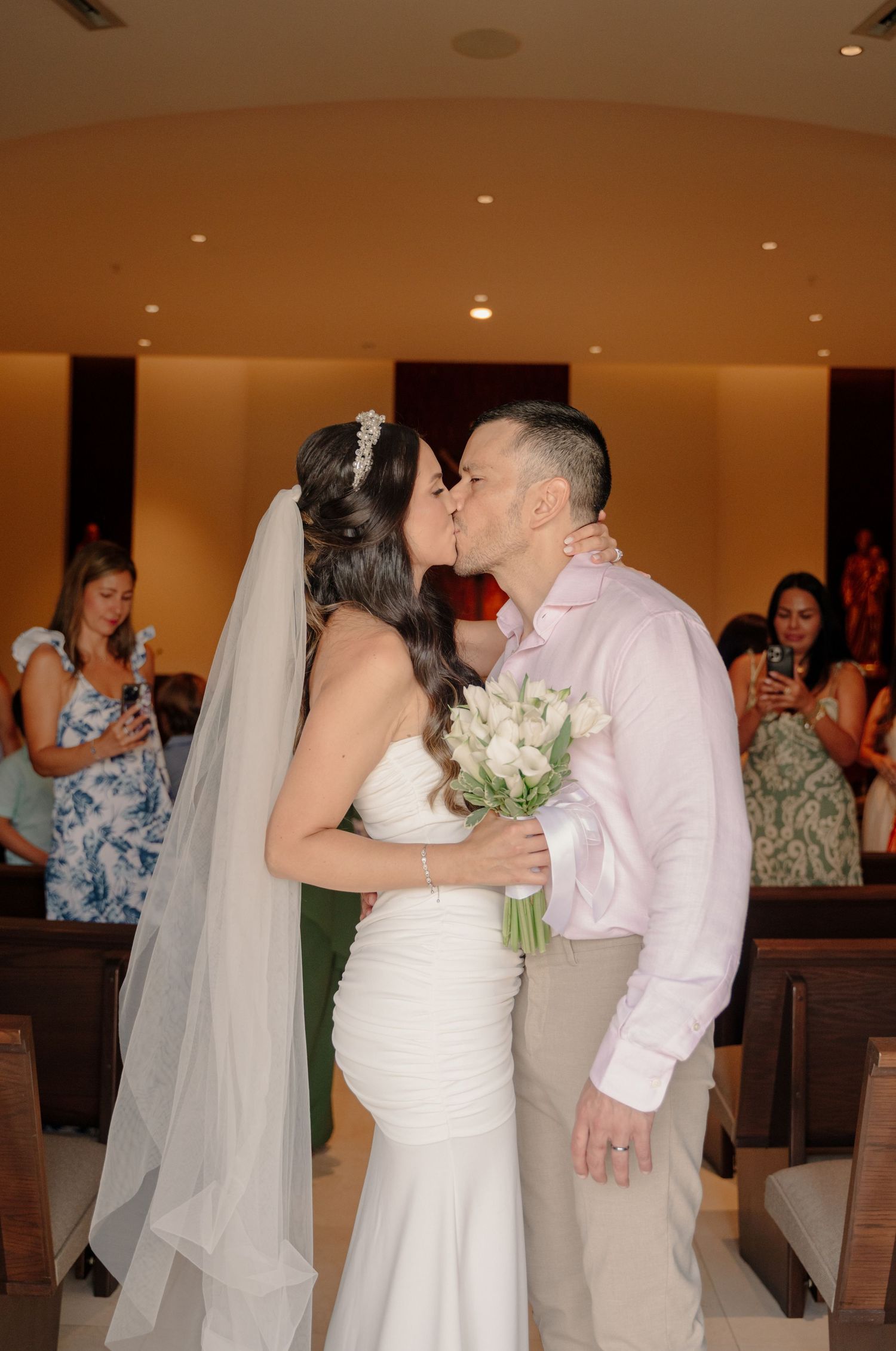 A romantic wedding moment captured as a couple shares a kiss while holding a white floral bouquet at their ceremony.