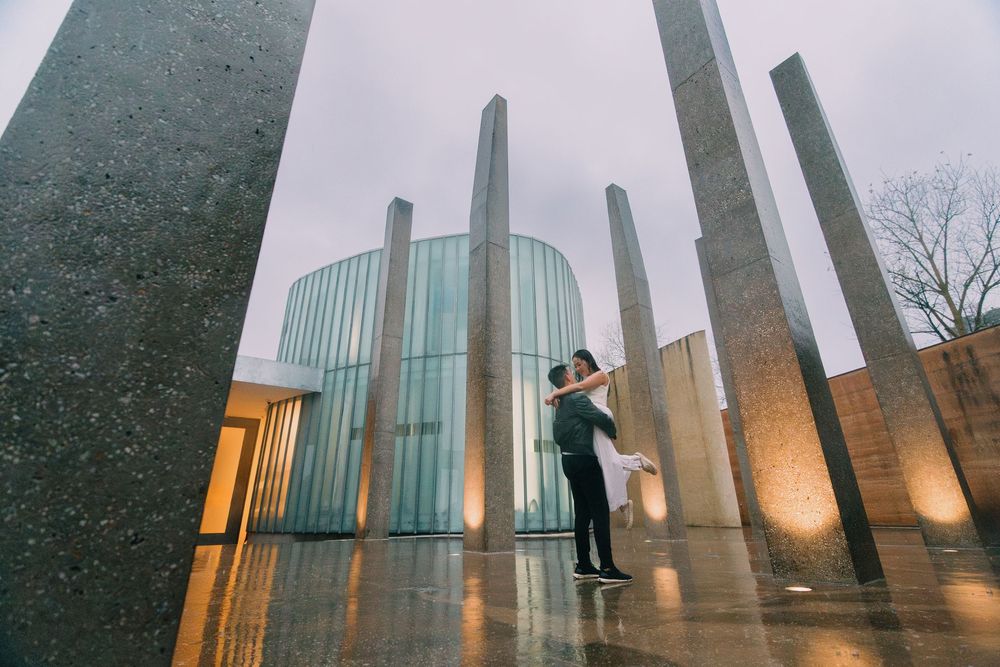 Two people share an intimate moment between tall modern concrete pillars at dusk.