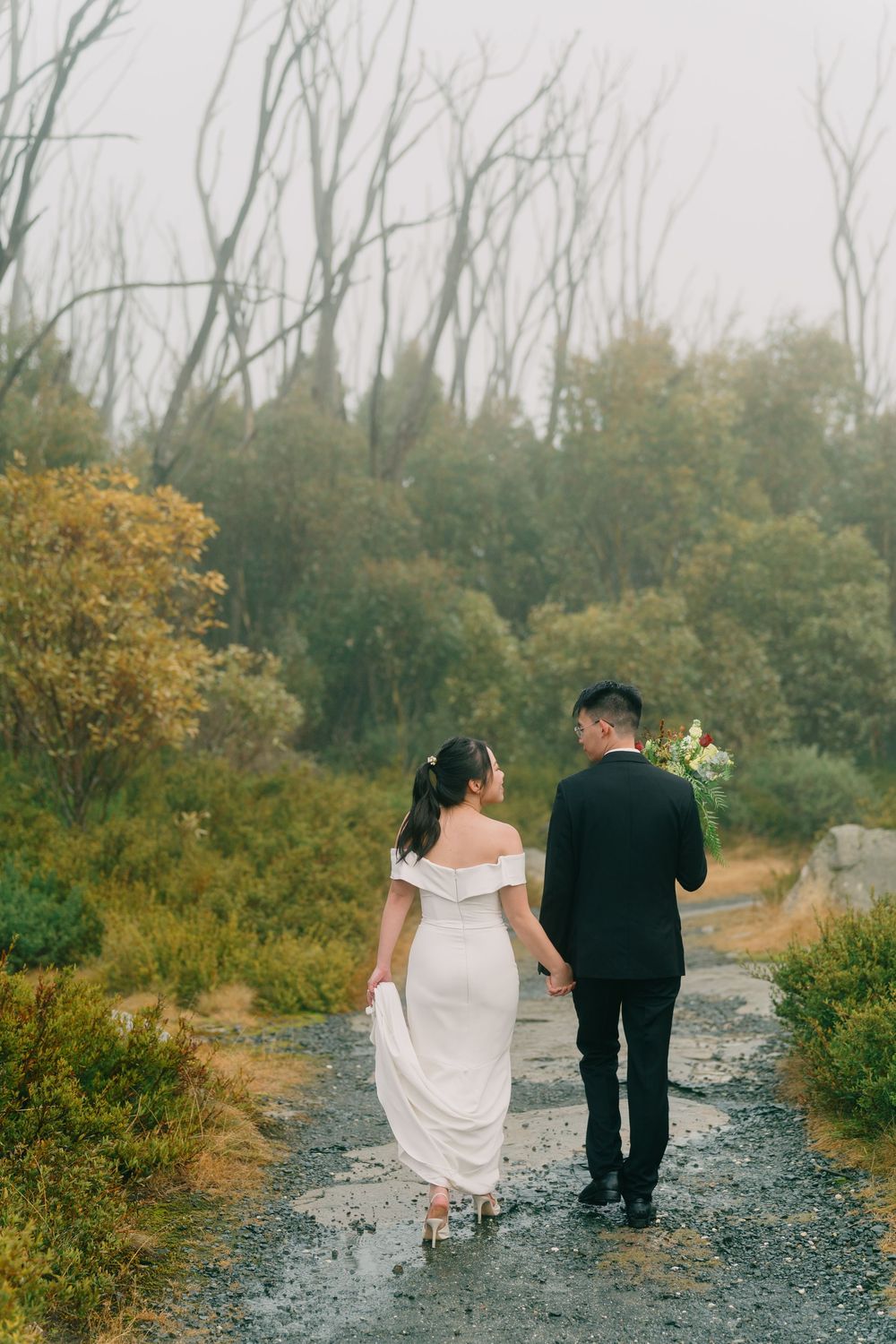 A couple walks down a gravel path surrounded by autumn foliage, the bride wearing a white dress and groom in black.