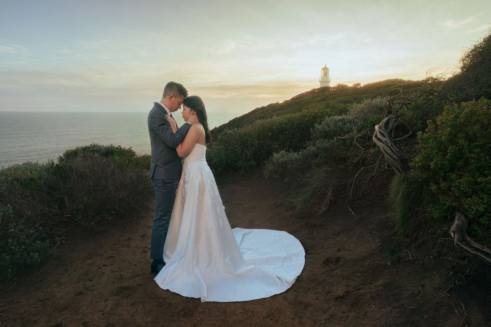 A romantic wedding couple embraces on a coastal cliff at sunset with a lighthouse in the distance.