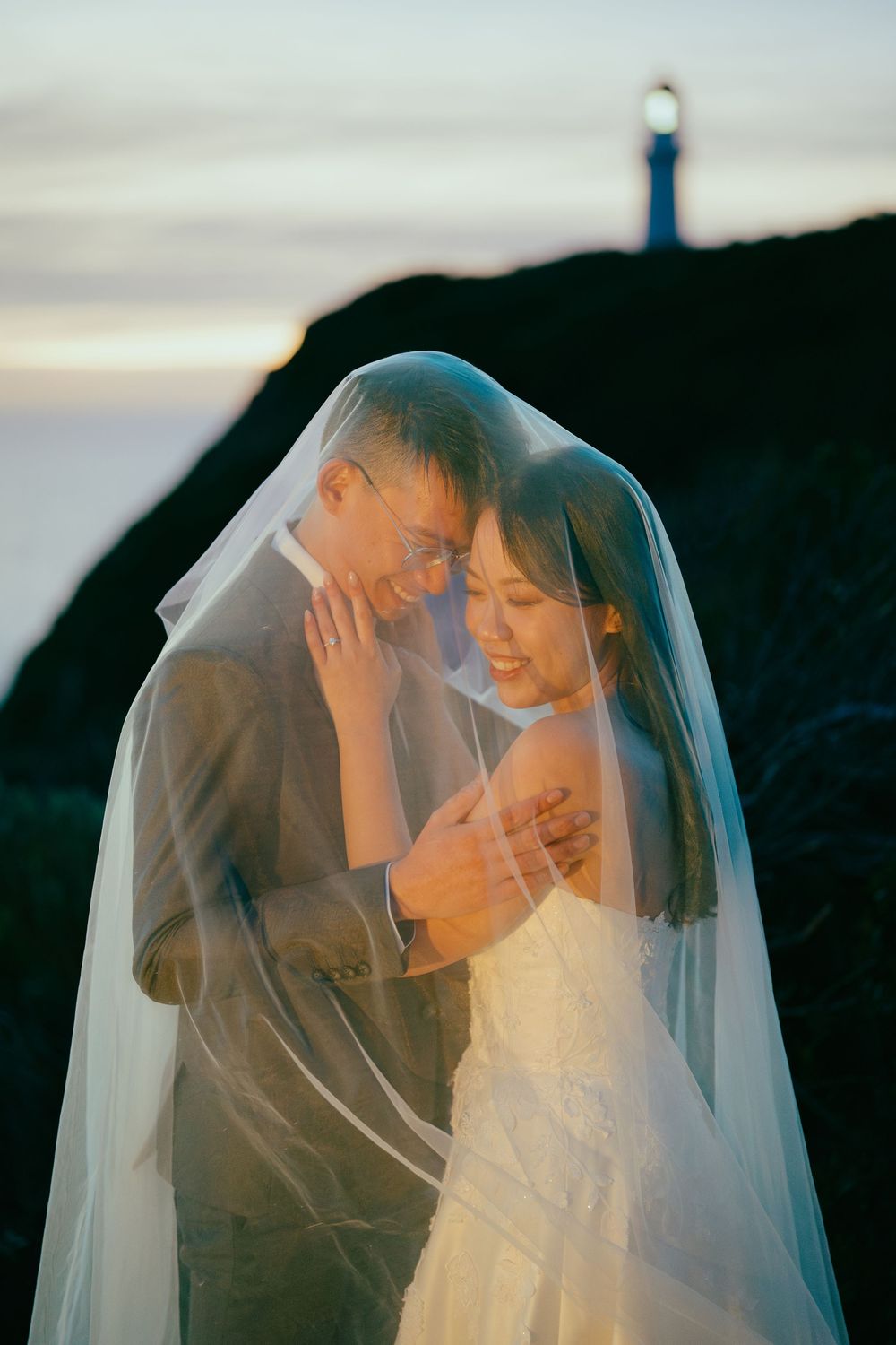 A newlywed couple shares an intimate romantic moment under a flowing white veil during sunset.