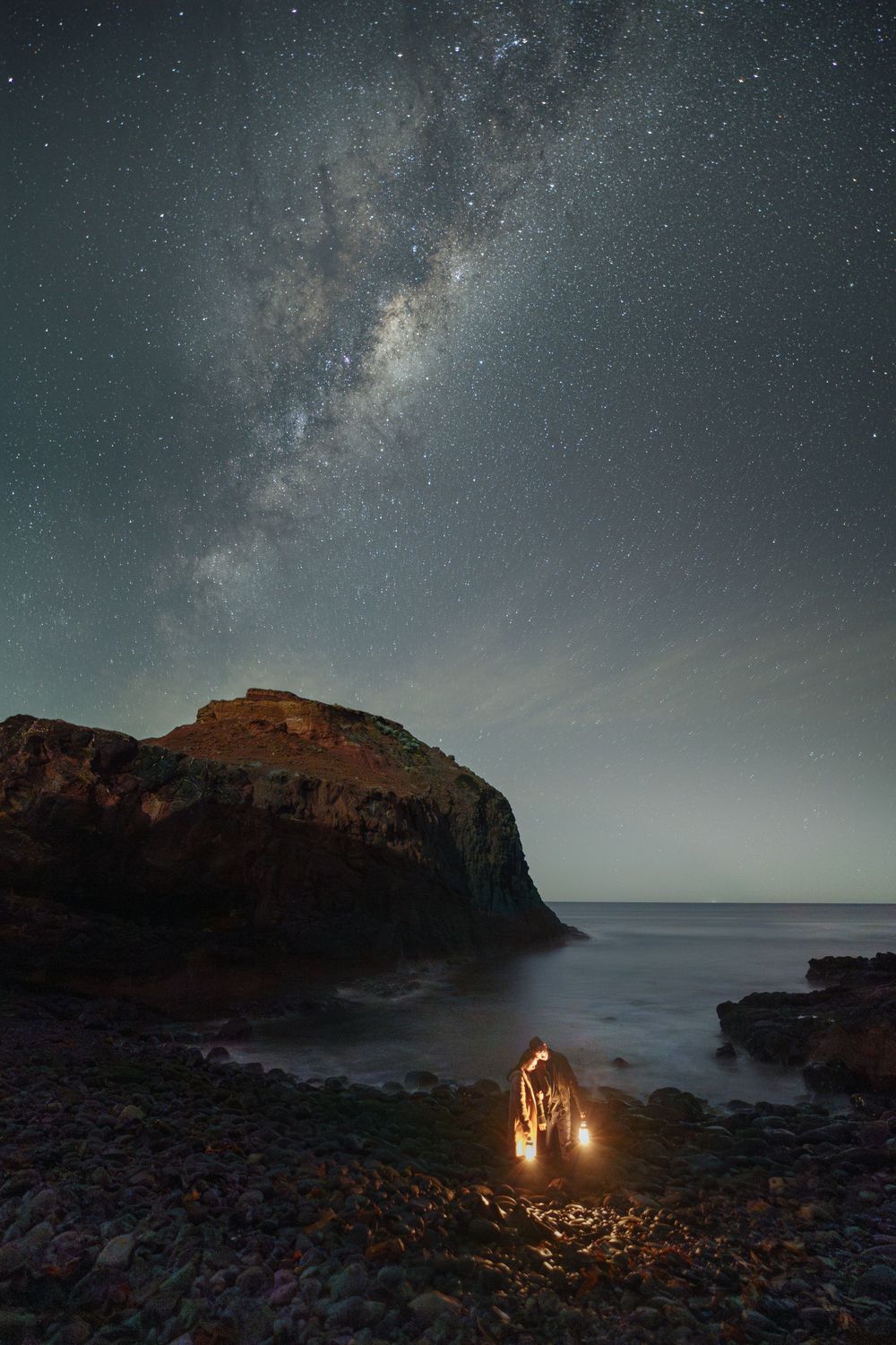 A panoramic sequence of the Milky Way galaxy stretching across a rocky coastal landscape with fire and torches glowing below.