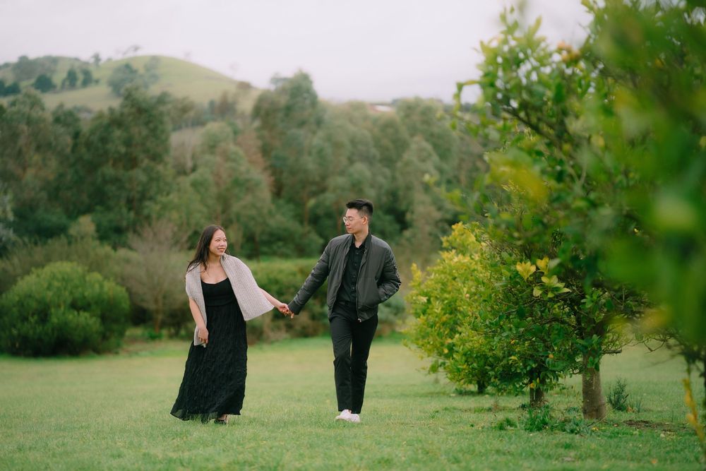 Couple walks together through serene meadow with lush greenery and rolling hills in the background.
