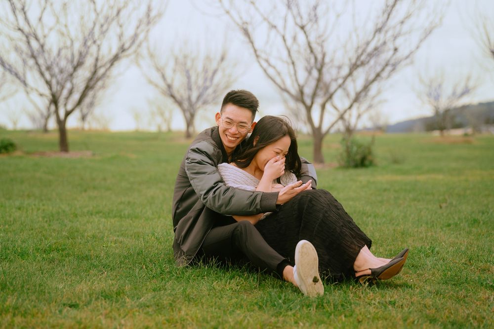 A couple sits together on green grass in an orchard during springtime, sharing a joyful moment together.