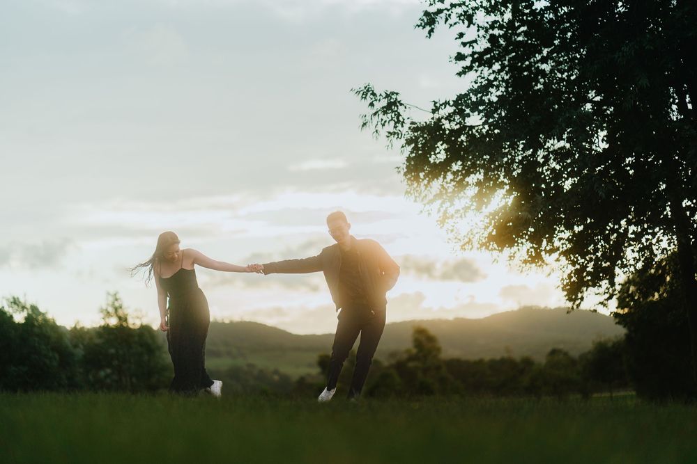 Silhouettes of two people holding hands and dancing in a grassy field at sunset with trees in background.