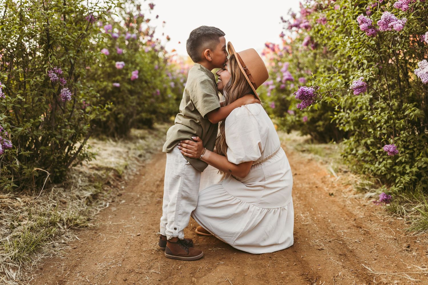 Parent and child embrace on a dirt path surrounded by blooming purple flowers and greenery in golden sunlight.